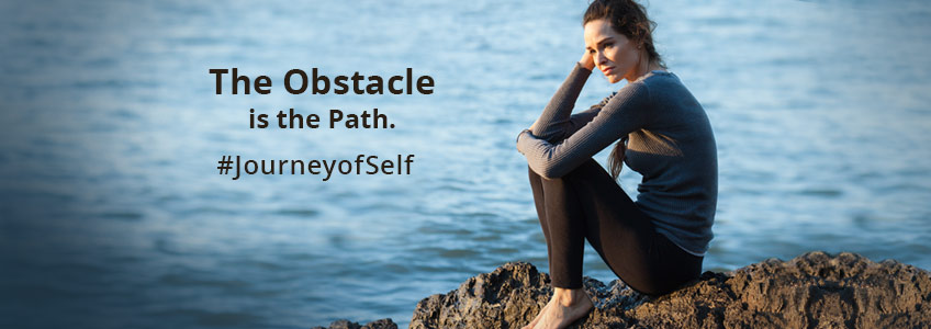 A thoughtful woman sitting on rocks by the water considers the obstacles in her path and how to overcome them as part of the Journey Of Self-Discovery. #JourneyofSelf