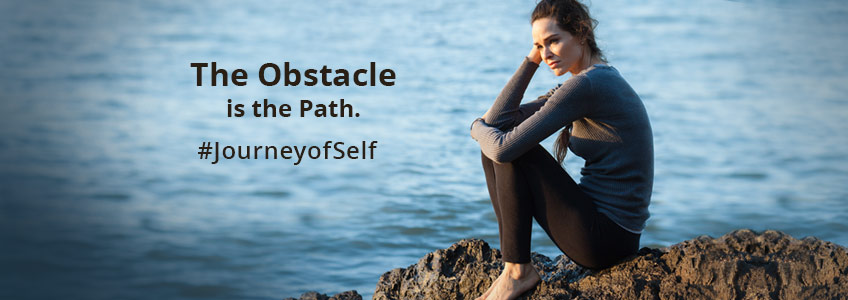 A thoughtful woman sitting on rocks by the water considers the obstacles in her path and how to overcome them as part of the Journey Of Self-Discovery. #JourneyofSelf